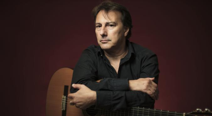 Mann mit dunklem Hemd hält eine klassische Gitarre, Arme verschränkt über dem Instrument, ernster Blick, vor dunkelrotem Hintergrund.