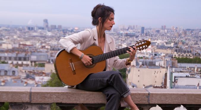 Frau mit hochgesteckten Haaren und weißer Bluse sitzt auf einer Steinbrüstung, spielt Gitarre mit Blick über eine Stadtlandschaft im Hintergrund.