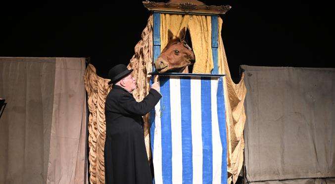 Mann in schwarzem Mantel und Zylinder füttert ein Pferd, das aus einer blauen Krippe mit weißen Streifen lugt, auf einer Bühne mit beigefarbenen Vorhängen.