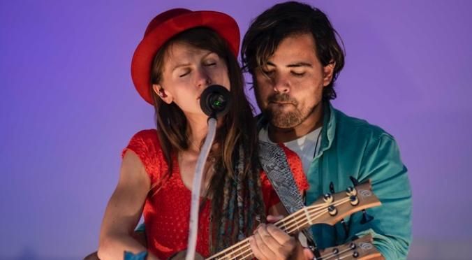 Zwei Musiker auf der Bühne: Frau mit rotem Hut singt ins Mikrofon, Mann mit Dreadlocks spielt Ukulele, beide konzentriert und nah beieinander.