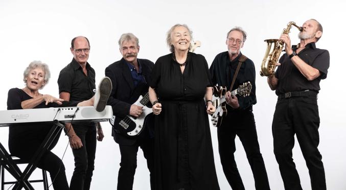 Sechs lächelnde Musiker: Frau am Keyboard, Mann mit hochgestelltem Fuß, zwei Gitarristen, Sängerin in schwarzem Kleid, Saxophonist – alle in schwarzer Kleidung vor weißem Hintergrund.