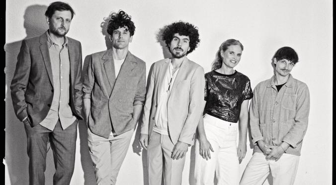 Fünf Personen in hellem Schwarz-Weiß-Foto: Vier Männer in Anzügen und Hemden, eine Frau in glitzerndem Oberteil und weißen Hosen, alle stehend nebeneinander vor neutralem Hintergrund.