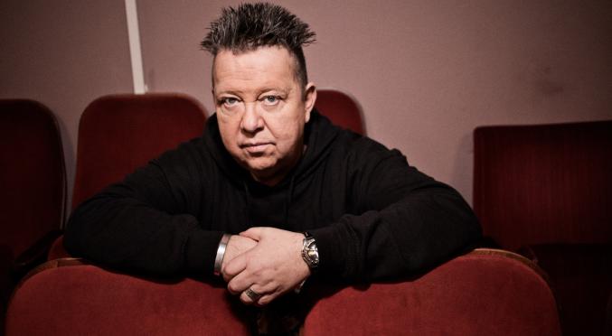 Mann mit aufrecht stehendem Haar und schwarzem Pullover sitzt nachdenklich mit verschränkten Armen auf roten Kinosesseln, Blick direkt in die Kamera.