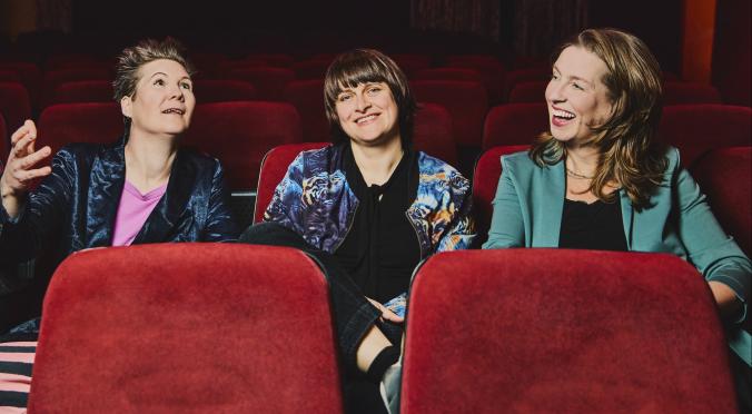 Drei lächelnde Personen sitzen in roten Kinosesseln: Links eine Frau mit kurzem Haar in glänzender Jacke, Mitte eine Person mit Pony in gemustertem Oberteil, rechts eine Frau mit langem Haar in grünem Blazer.