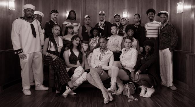 Gruppe von 18 Personen in maritimen und eleganten Outfits vor Holzvertäfelung: Matrosen, Offiziere und Personen in gestreifter Kleidung, stehend und hockend arrangiert.