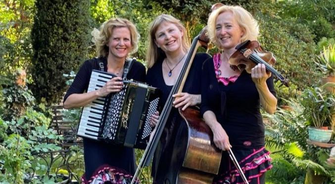 Drei lächelnde Frauen im Garten: Links spielt eine Akkordeon, Mitte hält einen Kontrabass, rechts spielt eine Geige, alle in schwarzen Outfits mit pinken Akzenten.
