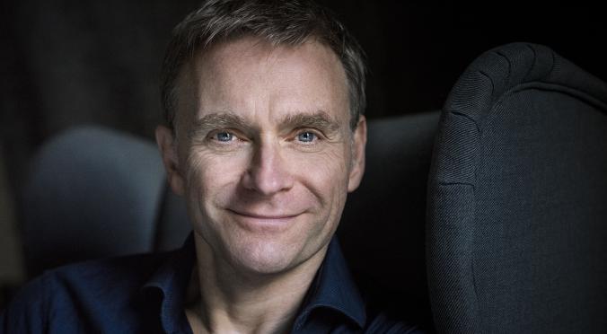 Lächelnder Mann mit kurzen graumelierten Haaren und blauem Hemd sitzt im Auto, die Hand auf der Kopfstütze des Sitzes, freundlicher Blick in die Kamera.