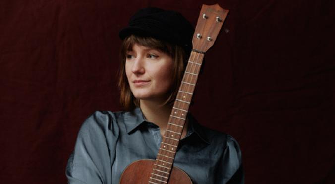 Lächelnde Person mit schwarzer Mütze und grauer Bluse hält eine braune Ukulele vor dunklem Hintergrund, Blick leicht zur Seite.