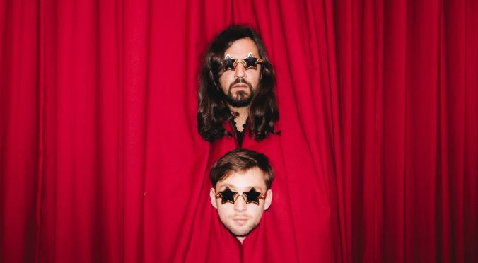 Zwei Personen stecken ihre Köpfe durch einen roten Bühnenvorhang: Beide tragen Sonnenbrillen in Sternenform, oben Person mit langen Haaren und Bart, unten Person mit kurzen Haaren.