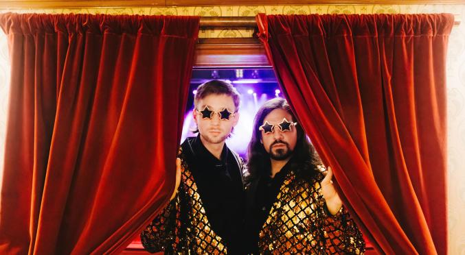 Zwei Personen mit glitzernden Jacken und Sternen-Sonnenbrillen posieren vor rotem Bühnenvorhang, eine hält den Vorhang auf, im Hintergrund blaue Bühnenbeleuchtung.