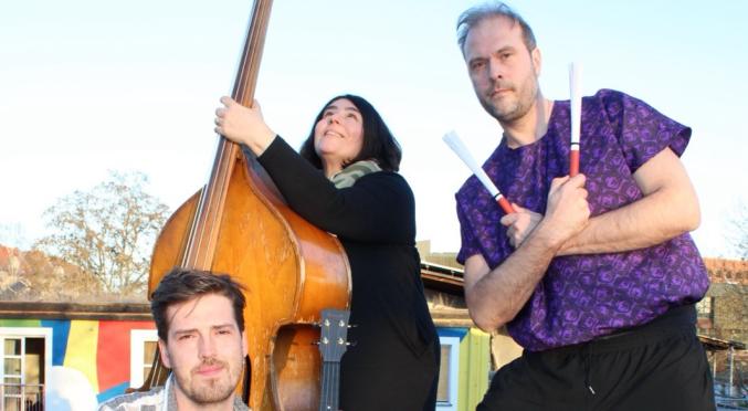 Drei Musiker:innen im Freien: Links sitzender Mann mit Ukulele, Mitte stehende Frau mit Kontrabass, rechts kniender Mann mit Stöcken vor einer Cajón-Kiste, bunte Häuser im Hintergrund.