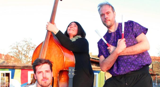 Drei Musiker:innen im Freien: Links sitzender Mann mit Ukulele, Mitte stehende Frau mit Kontrabass, rechts kniender Mann mit Stöcken vor einer Cajón-Kiste, bunte Häuser im Hintergrund.