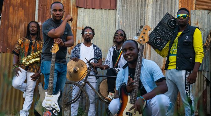 Sechs Musiker vor rostigem Wellblech: Links Saxophonist, Mitte Gitarrist, Bassist, Schlagzeuger mit Fahrradrahmen-Perkussion, rechts DJ mit Controller, alle lässig gekleidet, bunte Outfits.
