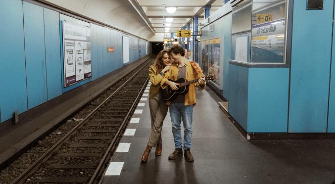 Zwei lächelnde Personen umarmen sich in einer U-Bahn-Station: Eine hält eine Gitarre, beide tragen lässige Kleidung, blaue Wandverkleidung und Gleise im Hintergrund.