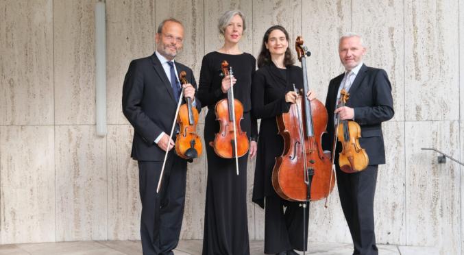 Vier Musiker:innen in schwarzer Kleidung vor hellem Steinmauer-Hintergrund: Zwei mit Violinen, eine mit Viola, einer mit Cello, alle Instrumente präsentierend.