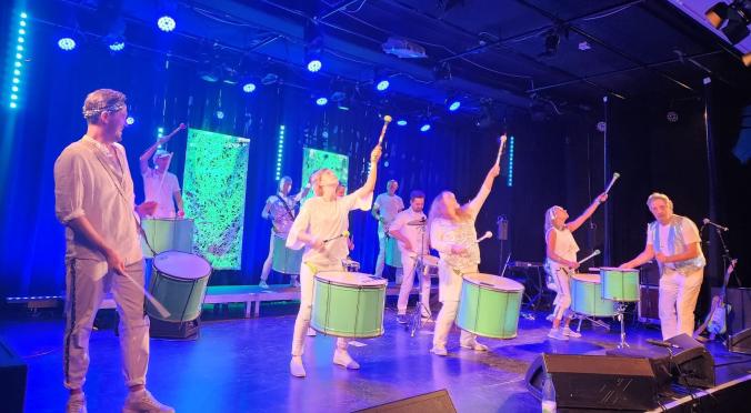 Sieben Musiker:innen in weißen Outfits spielen auf beleuchteter Bühne Trommeln und Perkussion, dynamische Posen, blaue Bühnenbeleuchtung und LED-Hintergrund.