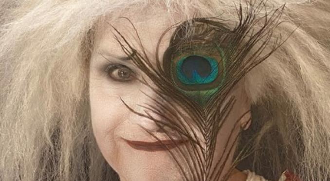 Person mit wilden, weißen Haaren und theatralischem Make-up hält einen Pfauenbusch vor ein Auge, trägt gemustertes, rotes Oberteil und weiße Ärmel.