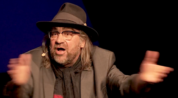 Mann mit Hut, Brille und rot-schwarz gestreiftem Schal steht mit ausgebreiteten Armen auf der Bühne, überraschter oder begeisterter Gesichtsausdruck, dunkler Hintergrund.