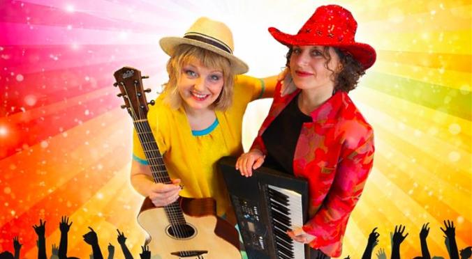 Zwei lächelnde Musikerinnen auf der Bühne: Links Frau mit Strohhut und Gitarre in gelbem Oberteil, rechts Frau mit rotem Hut und Keyboards, jubelndes Publikum und Regenbogen-Lichtstrahlen im Hintergrund.