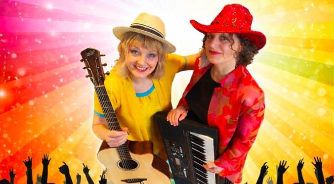 Zwei lächelnde Musikerinnen auf der Bühne: Links Frau mit Strohhut und Gitarre in gelbem Oberteil, rechts Frau mit rotem Hut und Keyboards, jubelndes Publikum und Regenbogen-Lichtstrahlen im Hintergrund.
