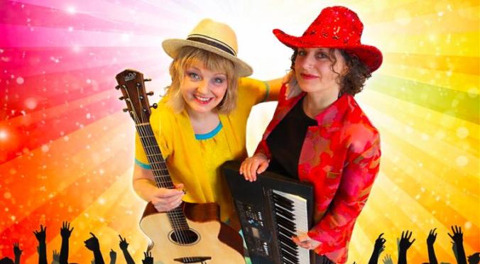 Zwei lächelnde Musikerinnen auf der Bühne: Links Frau mit Strohhut und Gitarre in gelbem Oberteil, rechts Frau mit rotem Hut und Keyboards, jubelndes Publikum und Regenbogen-Lichtstrahlen im Hintergrund.