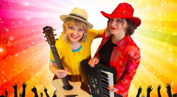 Zwei lächelnde Musikerinnen auf der Bühne: Links Frau mit Strohhut und Gitarre in gelbem Oberteil, rechts Frau mit rotem Hut und Keyboards, jubelndes Publikum und Regenbogen-Lichtstrahlen im Hintergrund.