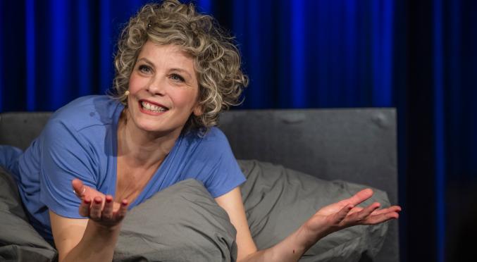 Lächelnde Person mit lockigen Haaren liegt entspannt in grauem Pyjama auf einem Bett, blaue Bettdecke, Arme ausgestreckt, als würde sie eine Geschichte erzählen, blauer Vorhang im Hintergrund.