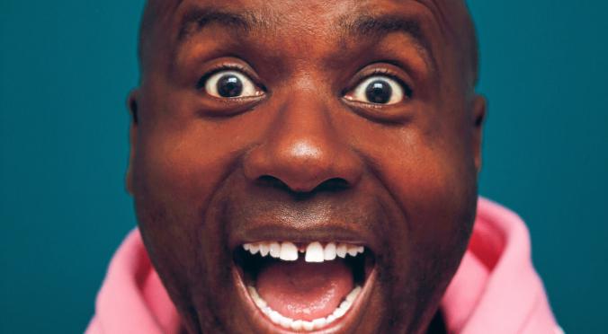 Lachelnde Person mit Glatze, weit geöffneten Augen und Mund, trägt einen rosa Kapuzenpulli, dunkler Hintergrund.