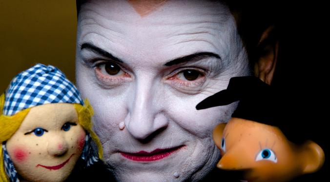 Person mit weißem Clown-Makeup, schwarzem Augenstreif, hält zwei Puppen: Links eine mit blau-kariertem Kopftuch, rechts eine mit orangefarbenem Haar und großen Augen.