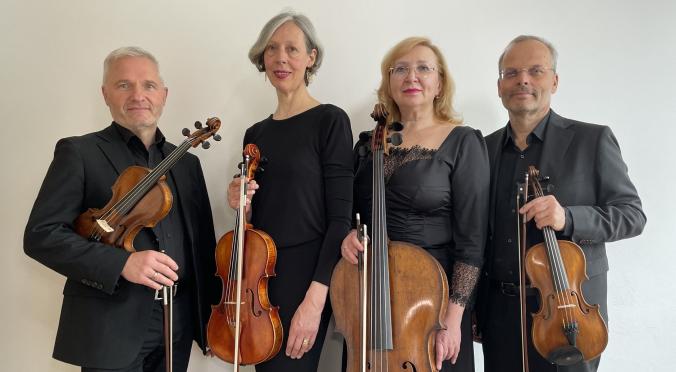 Vier lächelnde Musiker:innen in schwarzer Kleidung: Zwei mit Violinen, eine mit Cello, eine mit Bratsche, alle halten ihre Instrumente, helle Wand im Hintergrund.