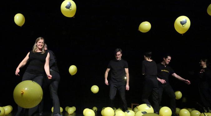 Fünf Personen tanzen auf dunkler Bühne zwischen vielen gelben Luftballons mit Hahnen-Aufdruck, alle in schwarzer Kleidung, dynamische Posen, fröhliche und lebendige Atmosphäre.