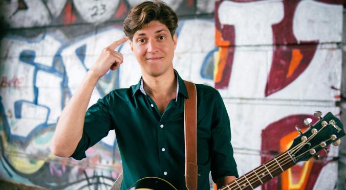 Lächelnder Mann mit dunklem Hemd und Gitarrenriemen hält eine schwarze Gitarre, posiert vor bunter Graffiti-Wand, Hand an der Schläfe, entspannte und kreative Atmosphäre.