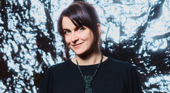 Lächelnde Person in schwarzem Pullover und Rock, mit glitzernder Kette als Krawattenersatz, steht vor funkelndem, beleuchtetem Hintergrund, Hände in den Taschen, kurze Haare.