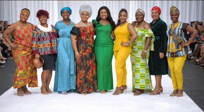 Sieben lächelnde Personen auf einem Laufsteg, alle in farbenfrohen, traditionellen afrikanischen Gewändern, verschiedene Muster und Farben, selbstbewusste Posen, Publikum im Hintergrund.