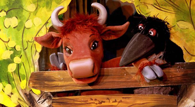 Stofffigur eines braunen Stiers mit großen Hörnern und freundlichem Gesicht lehnt über einem hölzernen Zaun, daneben eine schwarze Raben-Puppe, bunte Blumen im Hintergrund.