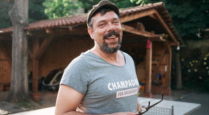 Mann mit Schiebermütze und T-Shirt hält seine Brille in der Hand und lacht und steht dabei vor einer Tischtennisplatte und einem Carport