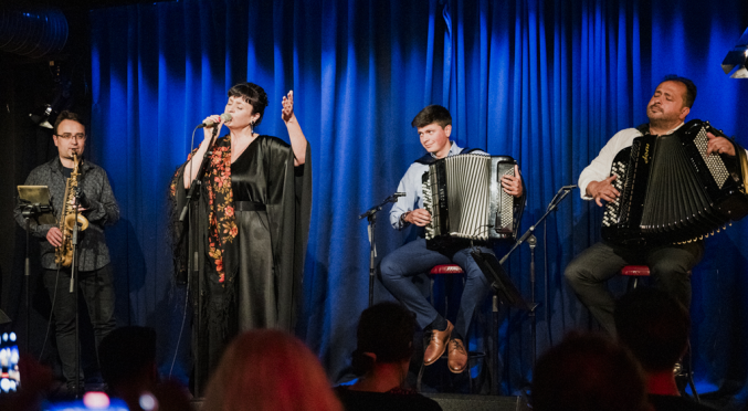 Vier Musiker auf der Bühne mit blauem Vorhang: Frau in schwarzem Kleid singt ins Mikrofon, links Saxophonist, rechts zwei Akkordeonspieler auf Hockern, Publikum im Vordergrund.