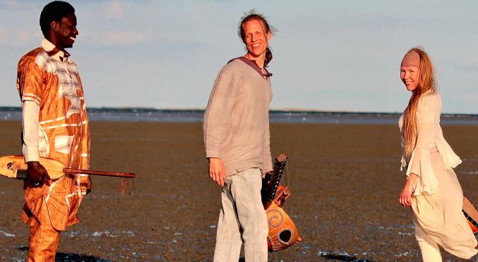 Drei Musiker am Strand: Links Mann mit traditionellem Instrument in orangefarbenem Gewand, Mitte Mann mit Gitarre in grauer Kleidung, rechts Frau mit Geige in weißem Kleid, Meer im Hintergrund.