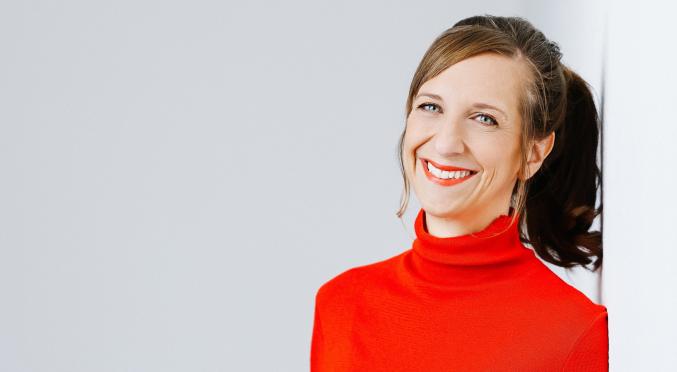 Frau mit Pferdeschwanz, rotem Rollkragenpullover und blauer Jeans lehnt lächelnd an weißer Wand, Hände in den Taschen, natürliches Licht.