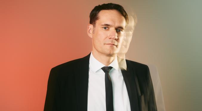Mann in dunklem Anzug, weißem Hemd und schwarzer Krawatte steht mit verschränkten Armen, ernster Blick, farbverlaufender Hintergrund von Orange zu Grün.