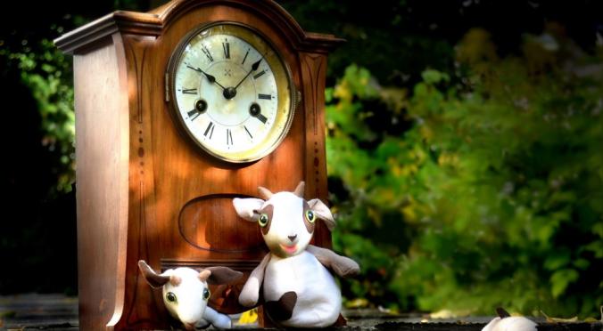 Sechs Stoff-Ziegenfiguren vor altem Holzwecker mit weißem Ziffernblatt, eine Ziege sitzt auf der Uhr, grüner Gartenhintergrund mit Blättern.