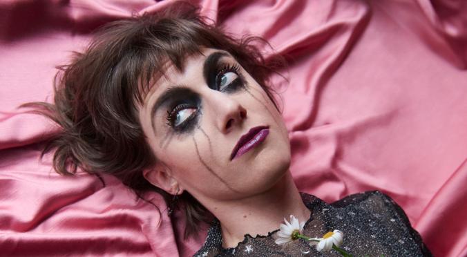 Frau mit dramatischem Make-up, schwarzem Sternenkleid und Gänseblumen im Haar liegt auf pinkem Satin, dunkles Haar, blauer Lidschatten, ernster Blick.