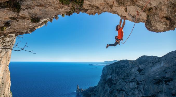 Person in orangefarbenem Shirt klettert an Felswand über türkisfarbenem Meer, Seil in der Hand, spektakuläre Aussicht auf Klippen und Horizont.