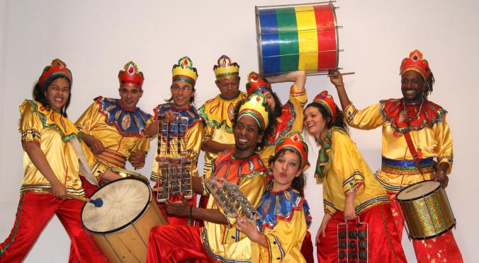 Acht Musiker in gold-rot-blauen Kostümen mit buntem Kopfschmuck, posierend mit Trommeln, Rasseln und einer grün-gelb-roten Trommel.