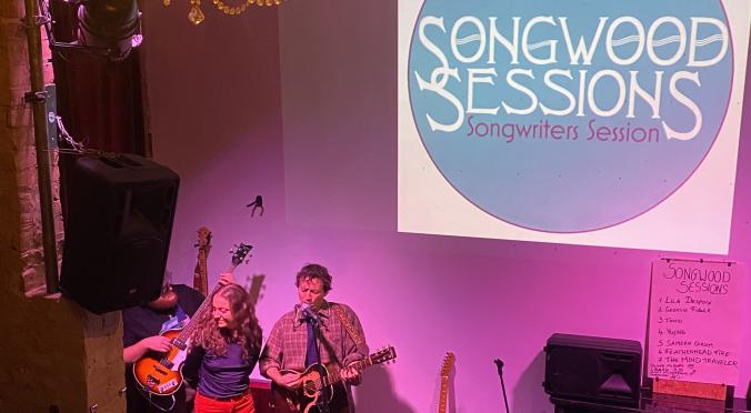 Live-Auftritt bei ‚Songwood Sessions‘: Drei Musiker mit Gitarre, Bass und Mikrofon auf kleiner Bühne, lila Beleuchtung, goldener Kronleuchter im Hintergrund.