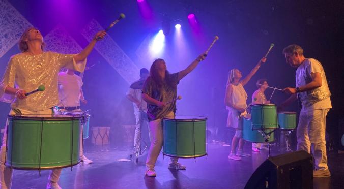 Sechs Musiker in weißen Outfits spielen grüne Trommeln auf einer pink beleuchteten Bühne, einige halten Schlägel in der Hand.