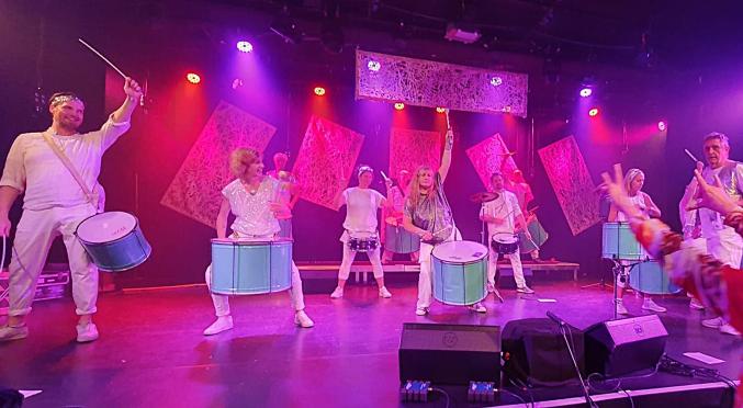 Sieben Musiker in weißen Outfits spielen Trommeln und Becken auf einer pink beleuchteten Bühne mit transparenten Vorhängen.