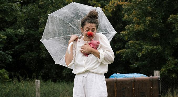Clown mit roter Nase hält durchsichtigen Regenschirm und pinken Ballon, trägt weißes Outfit, steht im Regen vor grünem Waldhintergrund.