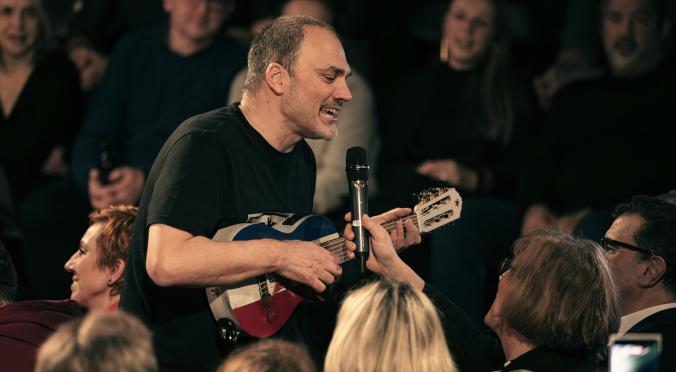 Mann in schwarzem Shirt spielt blaue Gitarre und singt ins Mikrofon, umgeben von aufmerksamen Zuhörern in dunklem Raum.