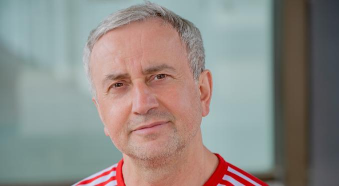 Mann mit graumeliertem Haar und Bart, trägt rotes Sportshirt mit weißen Streifen, vor hellem, unscharfem Hintergrund.